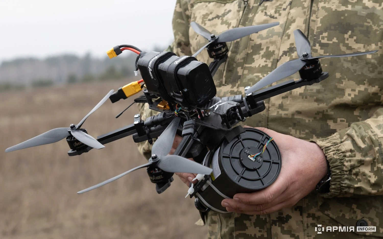 An FPV drone held by a person in military uniform, with propellers and motor visible, in an outdoor setting.