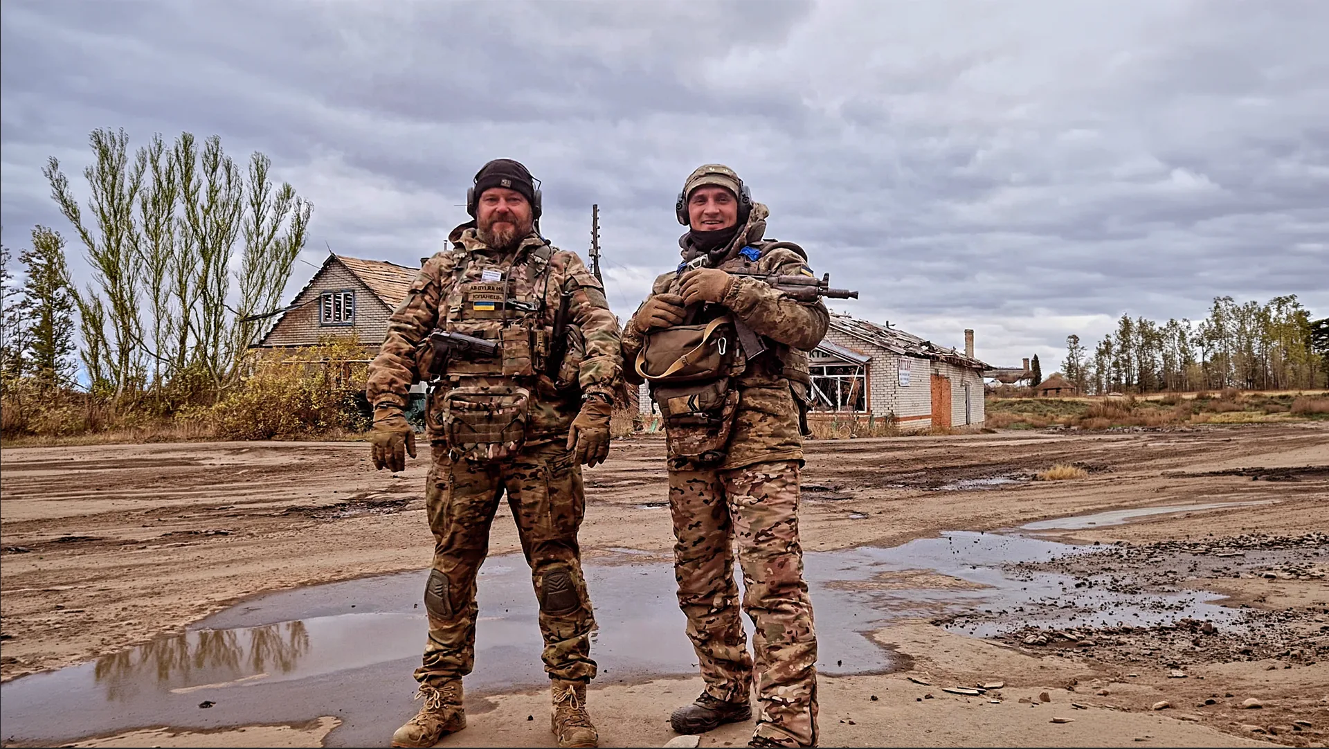 Deux hommes en tenue militaire se tiennent debout sur un terrain boueux, avec des bâtiments en arrière-plan sous un ciel nuageux.