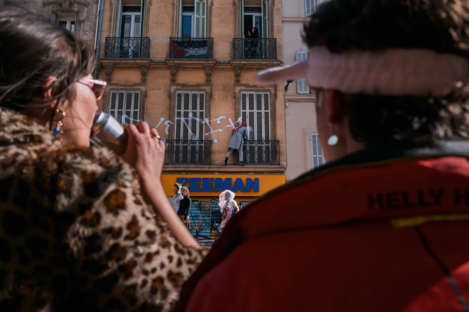 Deux personnes regardent un spectacle de carnaval, avec un participant sur un balcon et des décorations festives en arrière-plan.