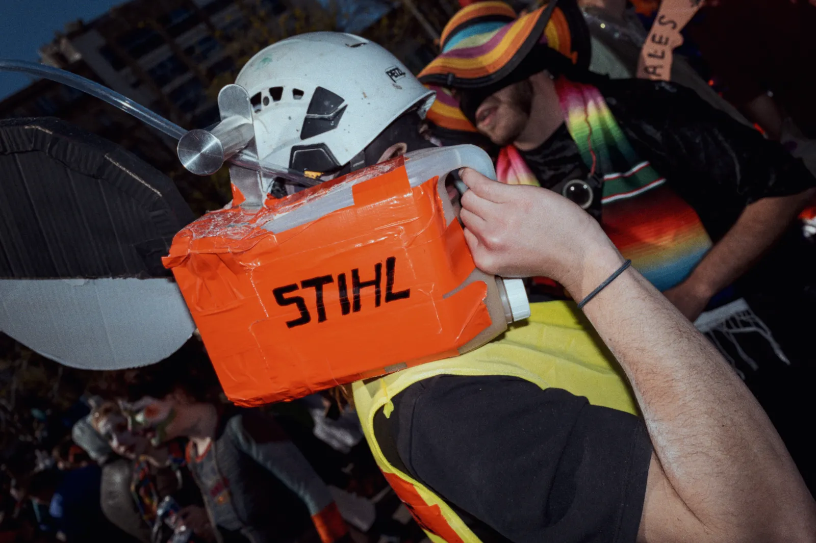 Personne portant un casque et un costume original, avec un accessoire en forme de tronçonneuse en carton marqué "STIHL". Ambiance festive.