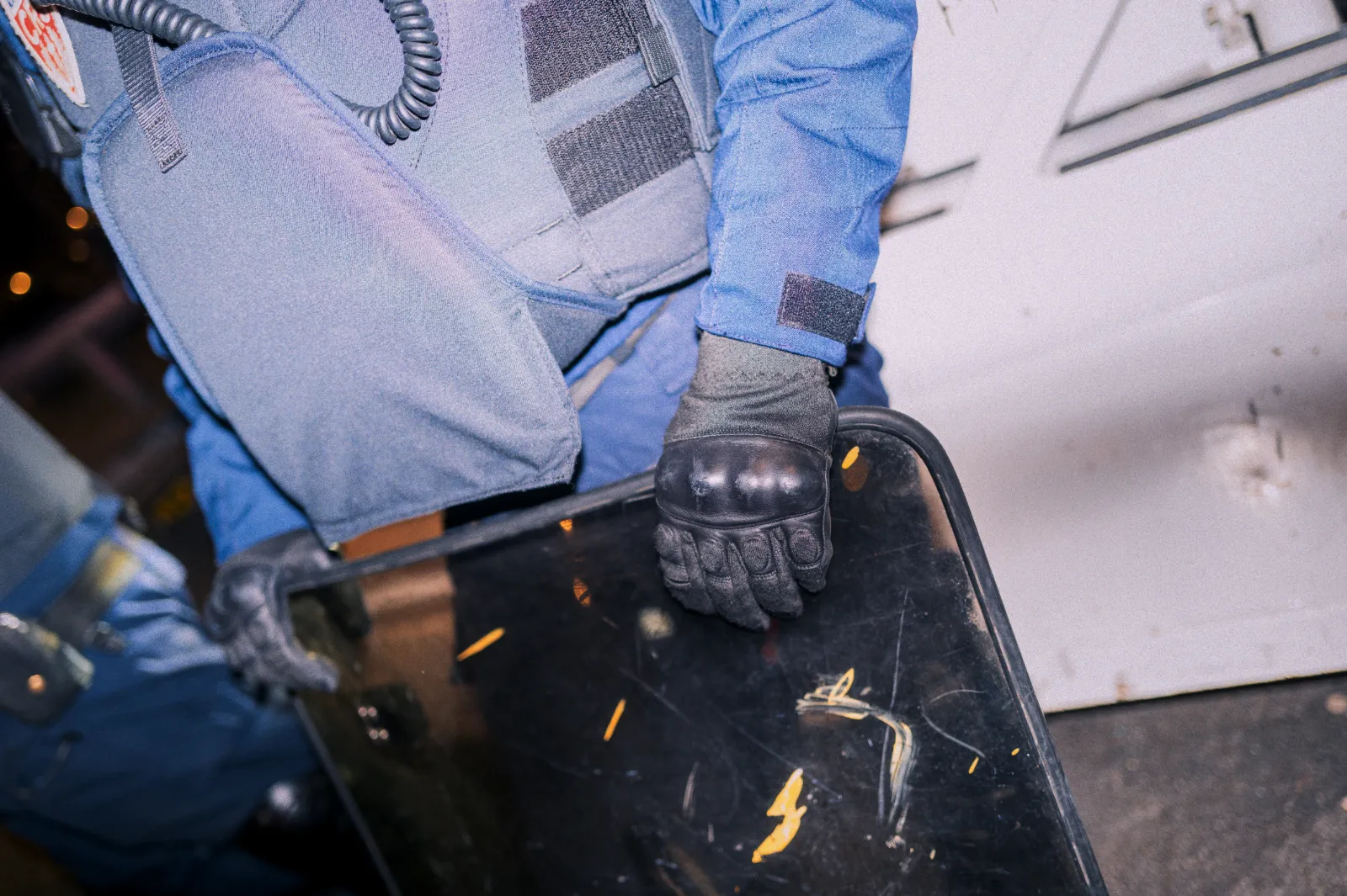 Un agent de sécurité tient un bouclier, vêtu d'un uniforme bleu et de gants noirs, dans un environnement urbain.