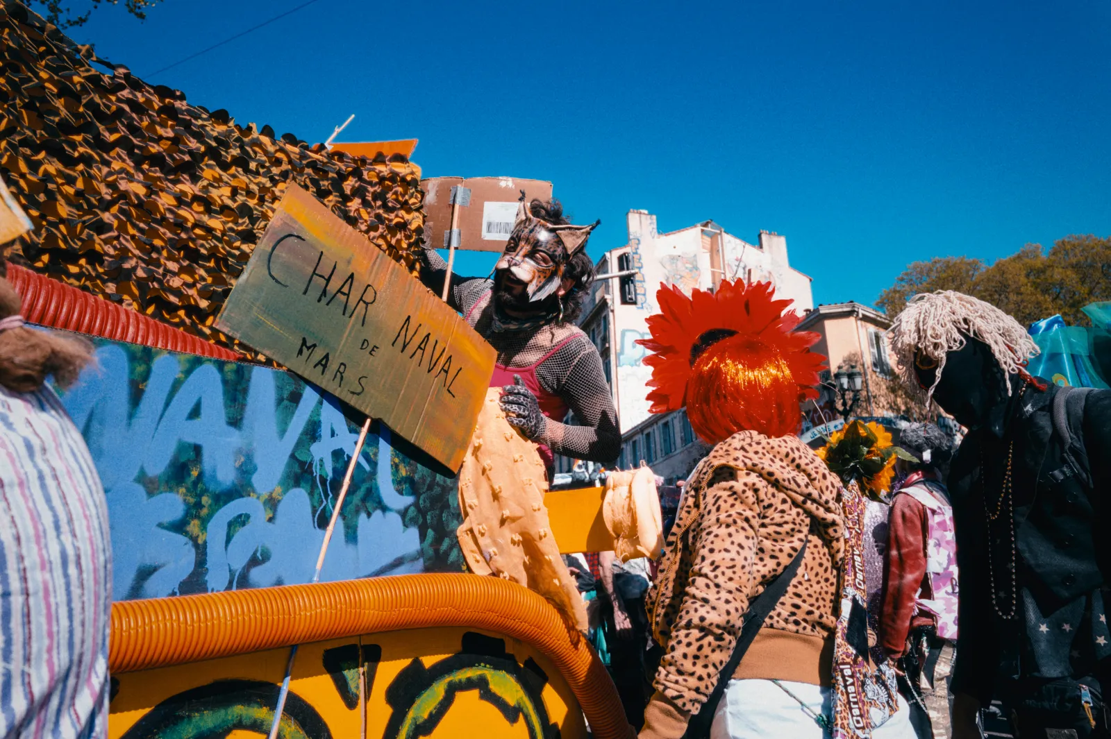 Des participants en costumes colorés autour d'un char décoré, avec un panneau "CHAR NAVAL de MARS", sous un ciel bleu.