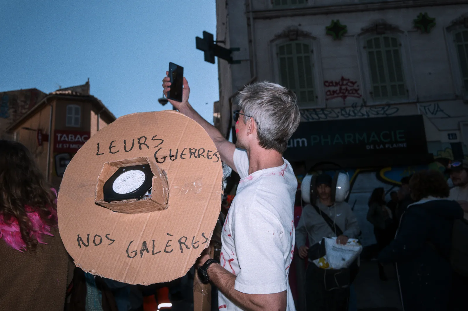 Un participant au carnaval tient un panneau en carton avec un message, tandis qu'il prend une photo avec son téléphone.