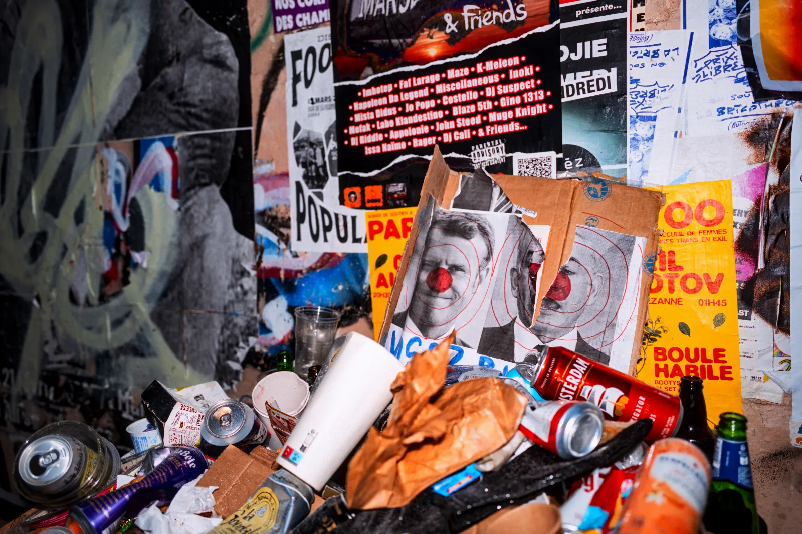 Débris et affiches collées sur un mur, avec des canettes, des papiers et des objets divers éparpillés au sol.
