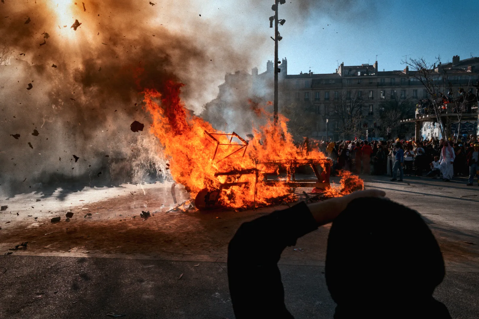 Flammes et débris s'élèvent d'un véhicule en feu lors du Carnaval de la Plaine, entouré de spectateurs.