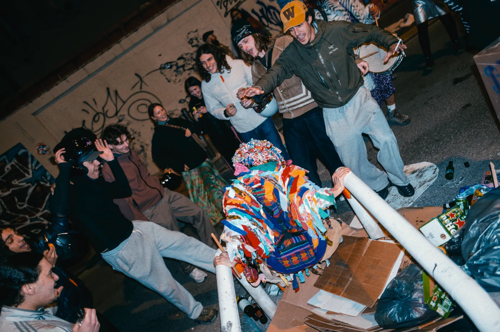 Groupe de personnes en fête autour d'une figure colorée, dans une ambiance festive et décontractée, avec des graffitis en arrière-plan.