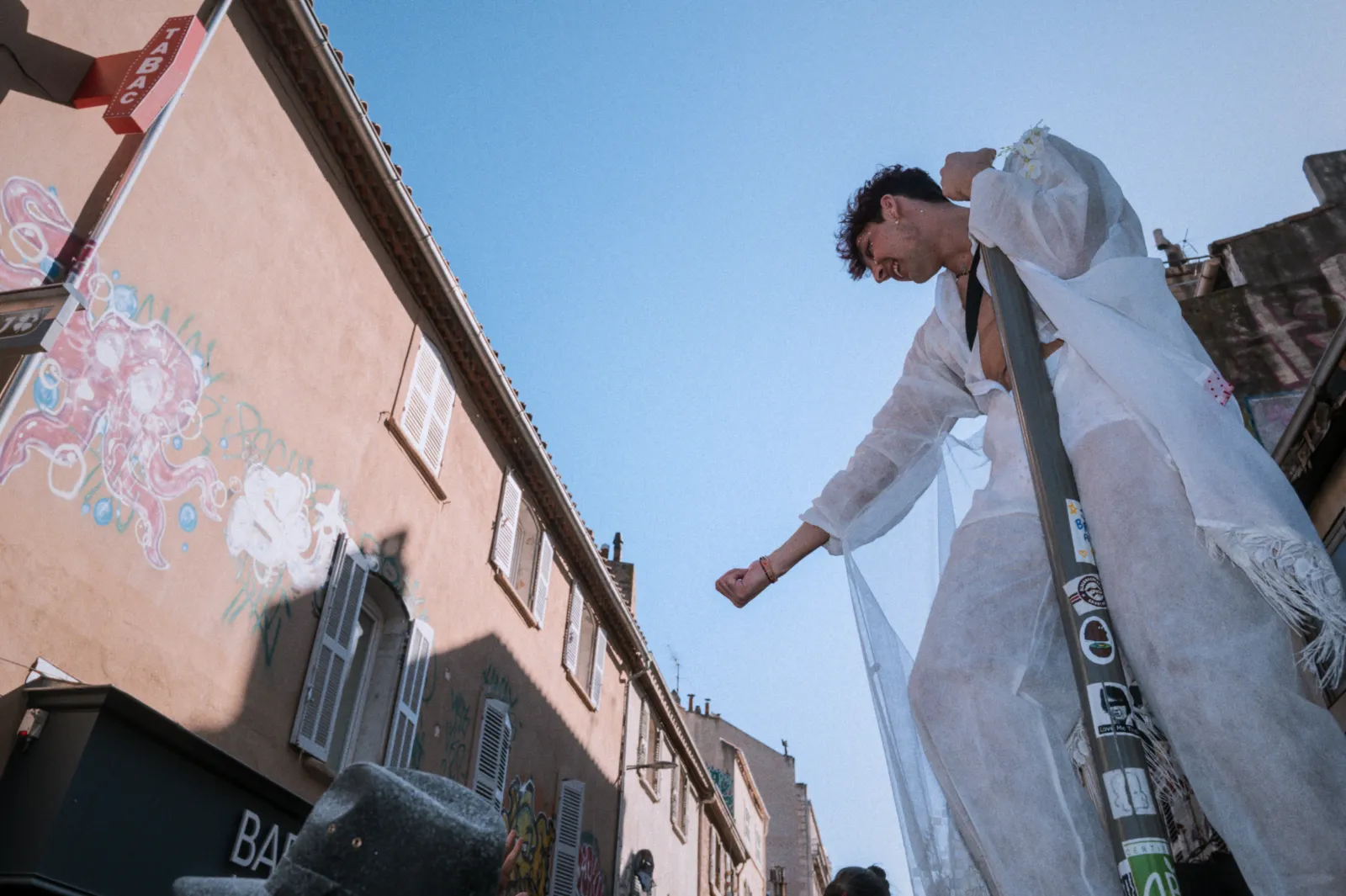 Un participant en costume festif danse sur un mât, sous un ciel bleu, avec des bâtiments colorés en arrière-plan.