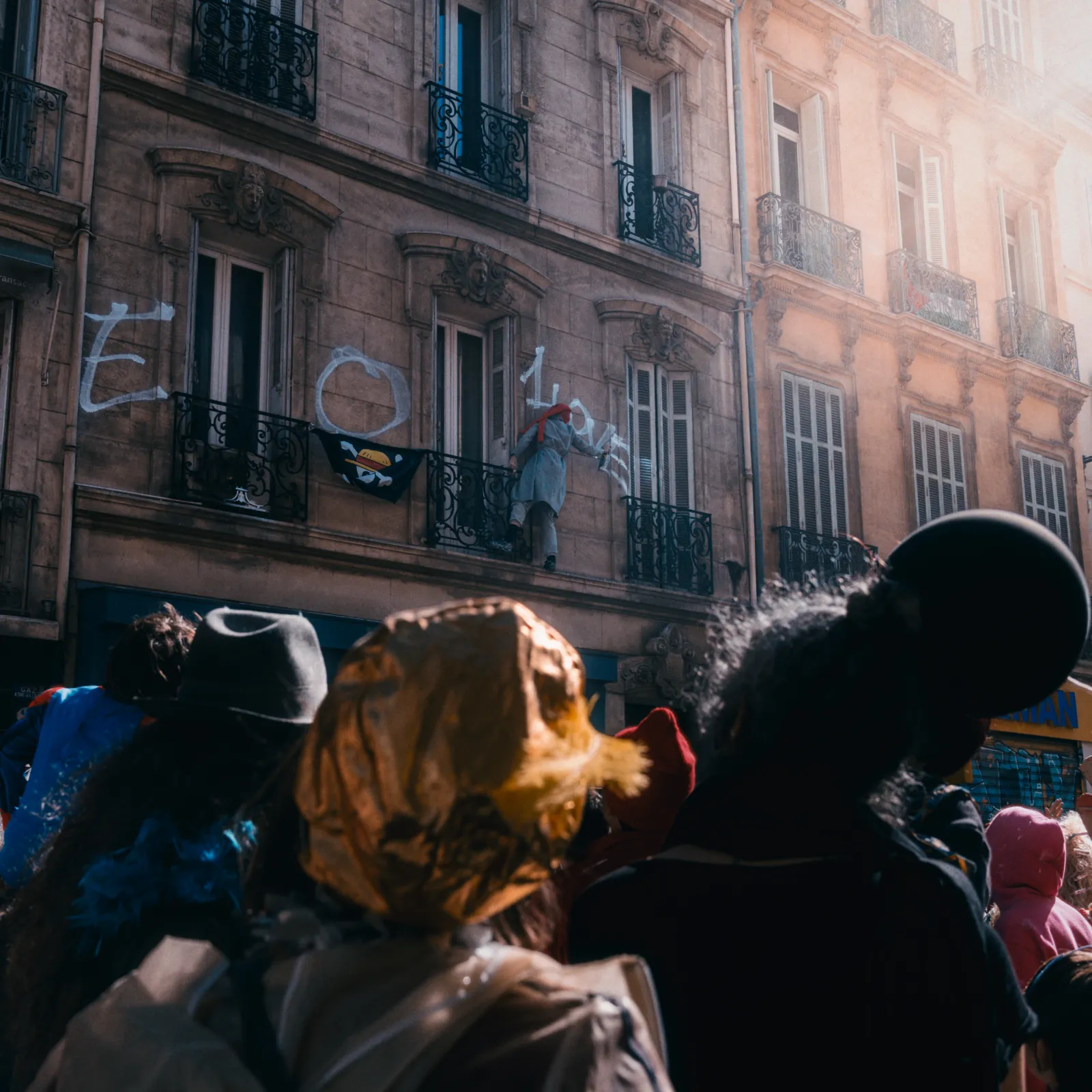 Une foule colorée regarde un personnage sur un balcon, avec des inscriptions sur le mur. Ambiance festive du carnaval.