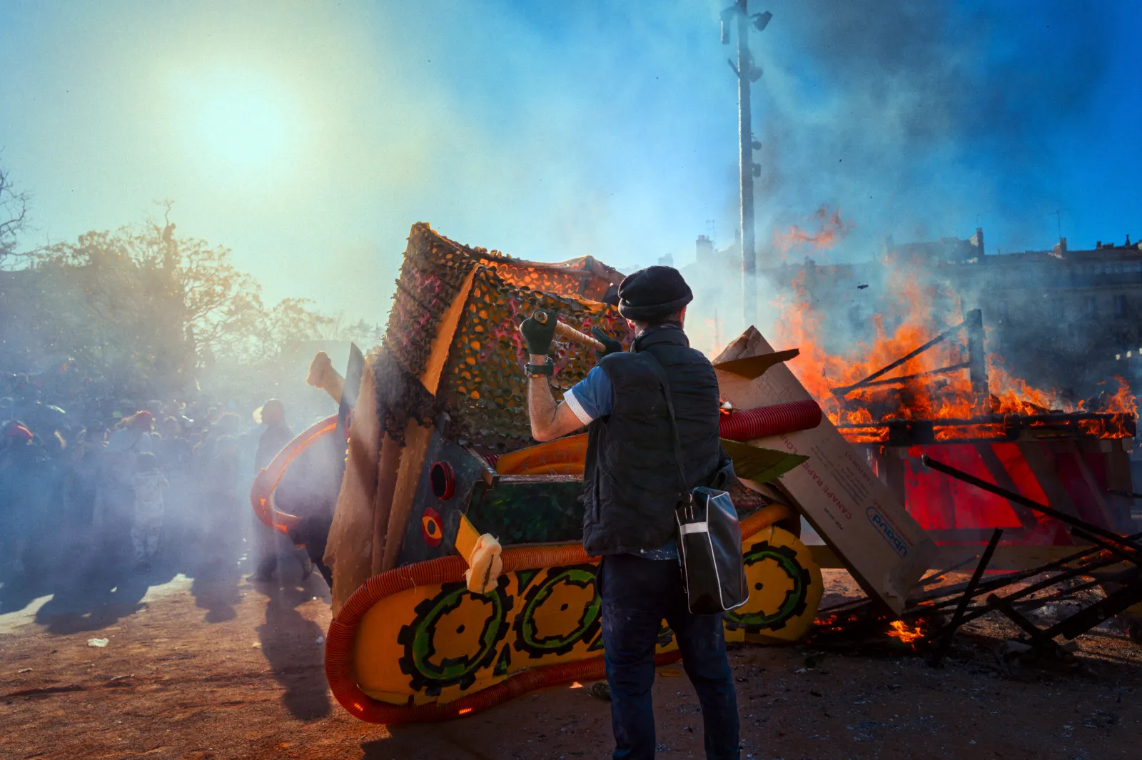 Un homme observe un char en feu au Carnaval de la Plaine, entouré de fumée et de spectateurs, sous un ciel ensoleillé.