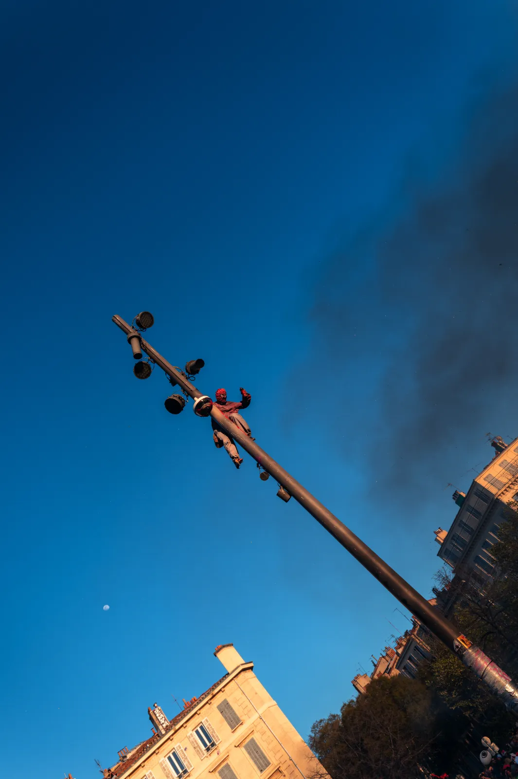 Un participant grimpe à un lampadaire sous un ciel bleu, avec des bâtiments en arrière-plan et une légère fumée.