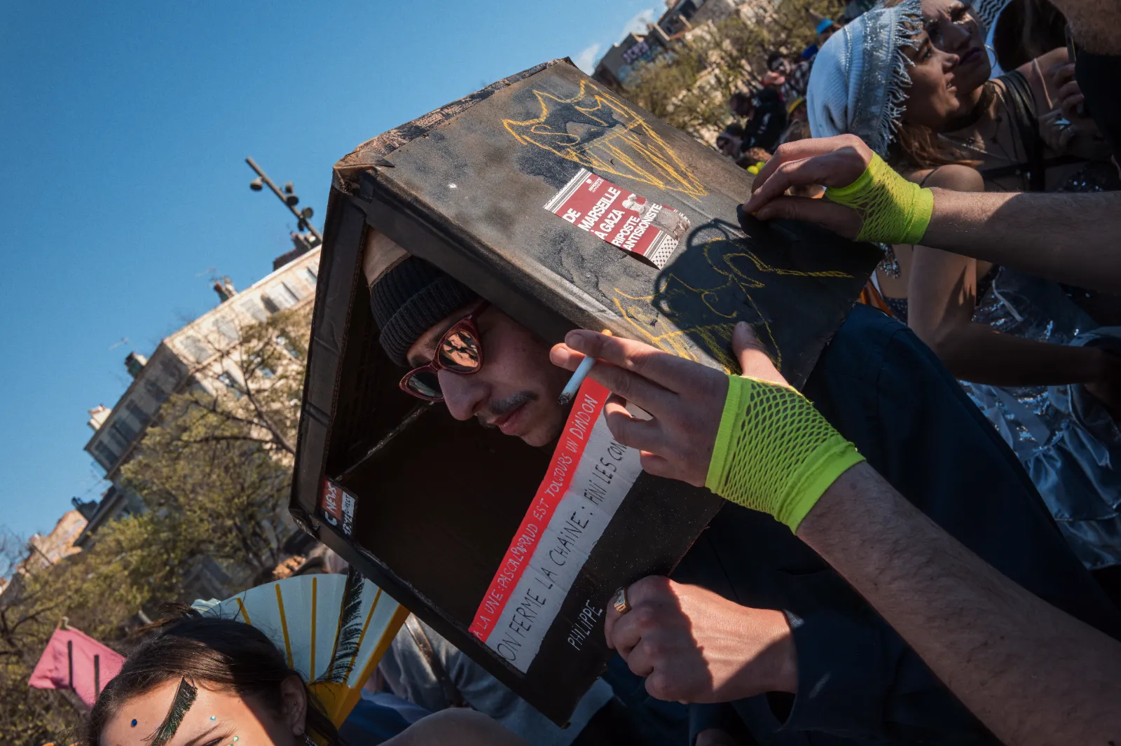Un participant au carnaval porte une boîte en carton sur la tête, entouré de personnes en costumes colorés sous un ciel bleu.