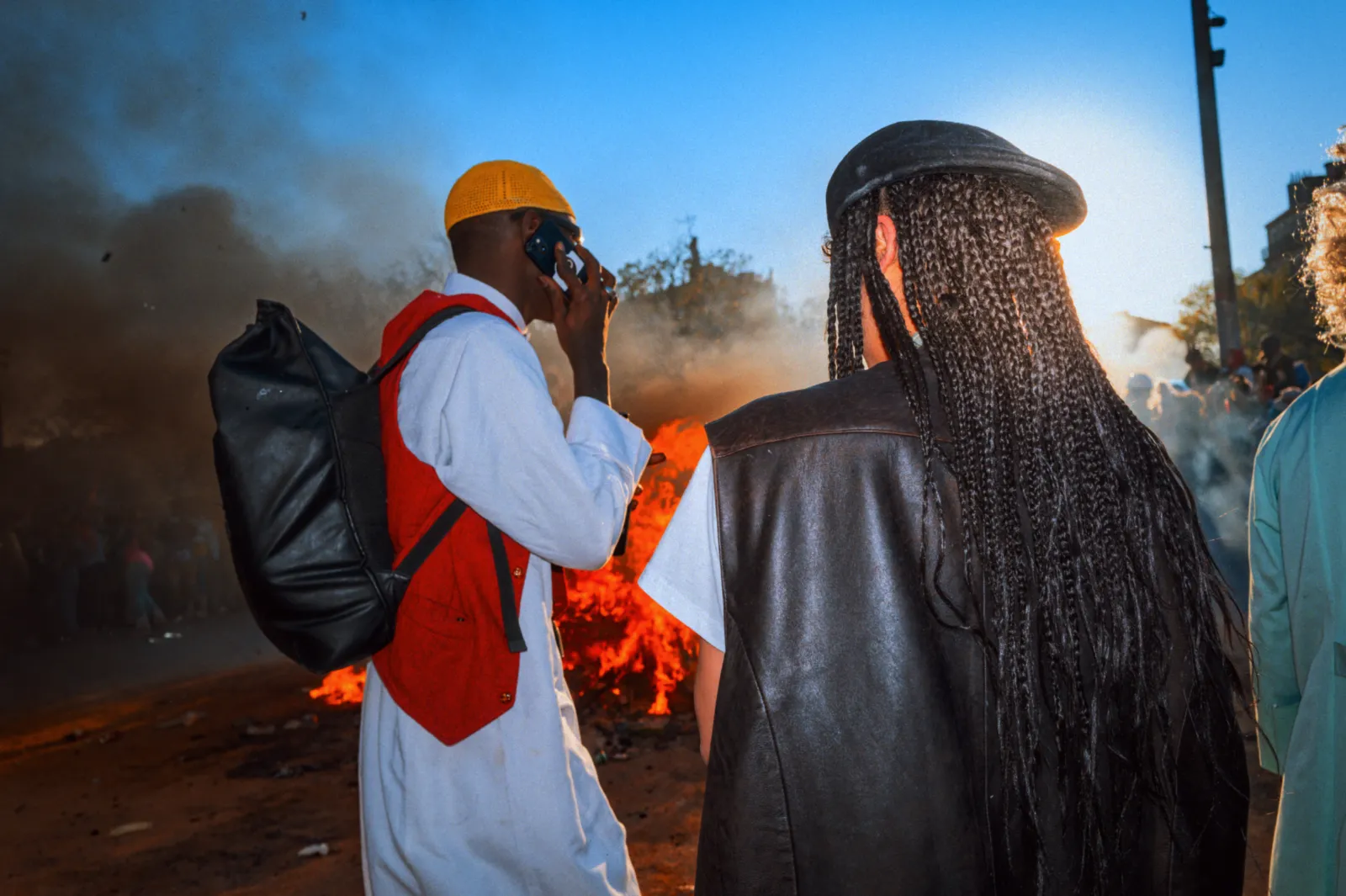 Deux personnes au carnaval, l'une portant un chapeau jaune et un sac à dos, l'autre avec des cheveux tressés, devant un feu.
