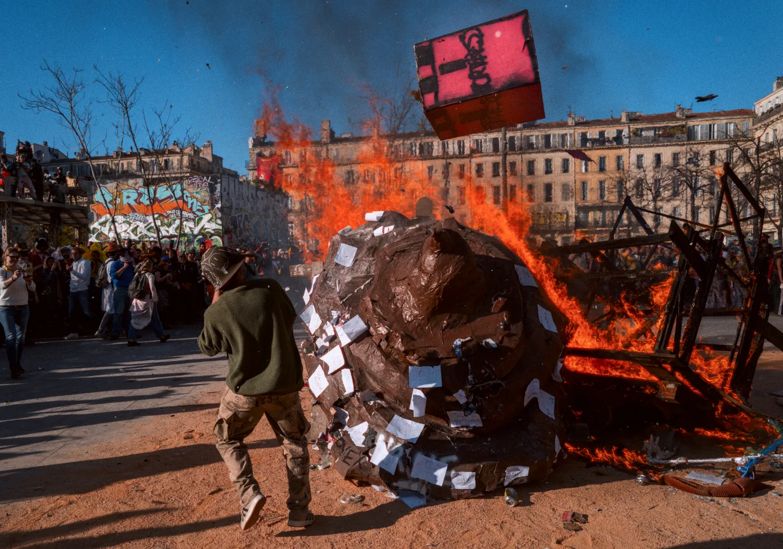 Un grand bonhomme en papier brûle au milieu d'une foule festive, avec des bâtiments colorés en arrière-plan et des graffitis.