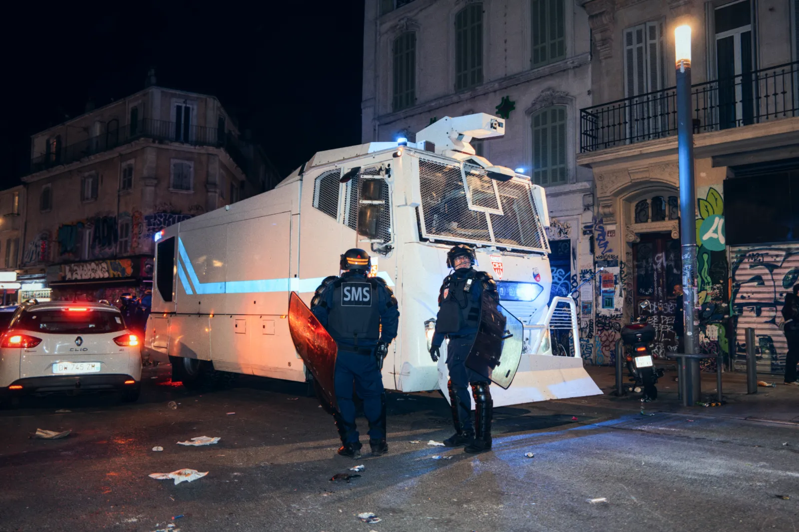 Véhicule de police blindé stationné dans une rue, avec des agents en tenue anti-émeute, graffiti en arrière-plan. Débris au sol.