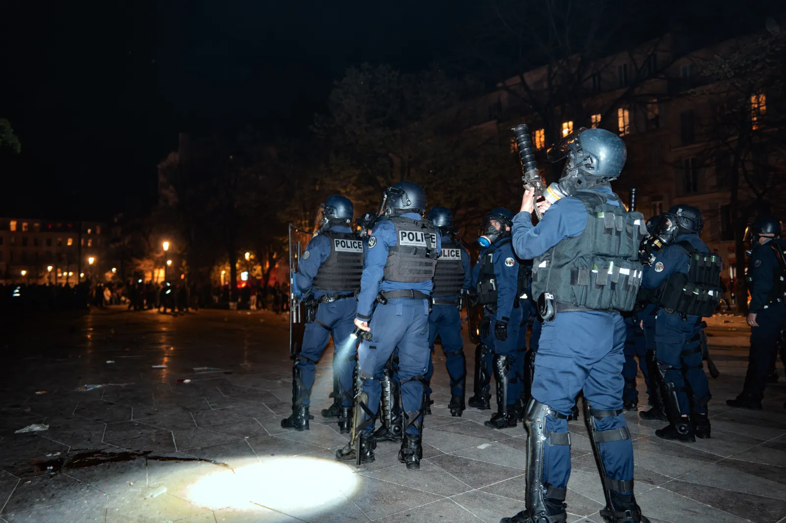 Des policiers en tenue anti-émeute surveillent une scène nocturne, équipés de lampes et d'armes, dans une ambiance de carnaval.