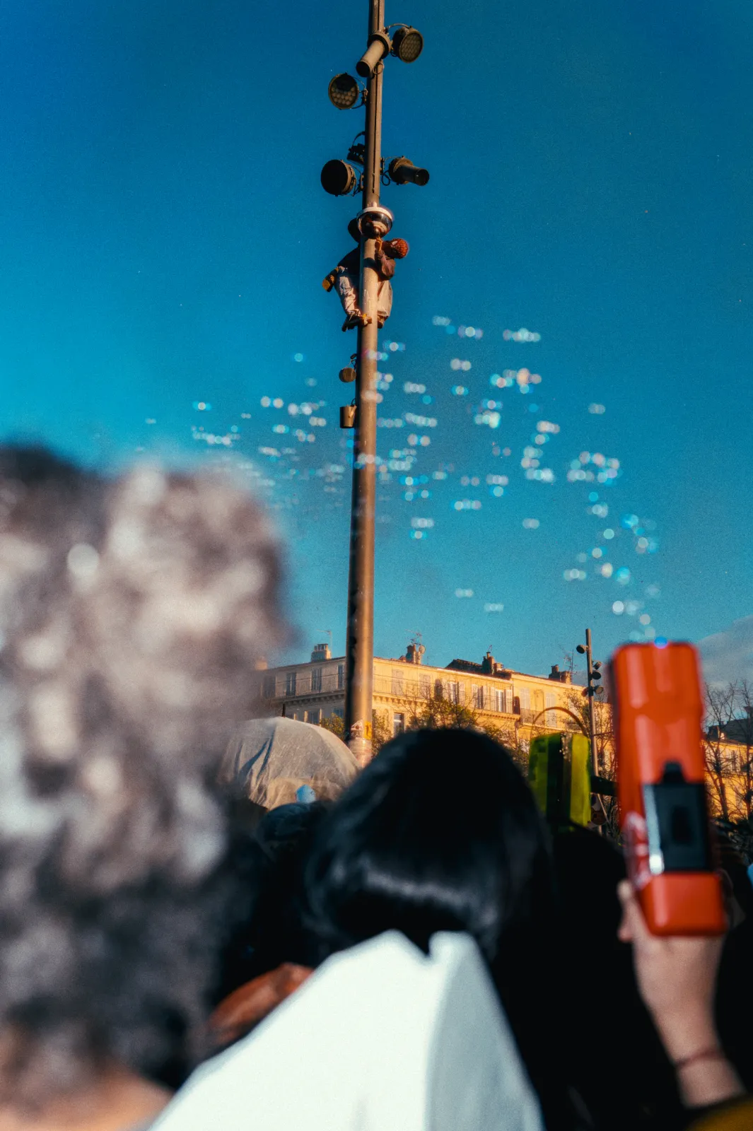 Des bulles flottent dans le ciel bleu au-dessus d'une foule festive, avec un lampadaire en arrière-plan et des bâtiments.
