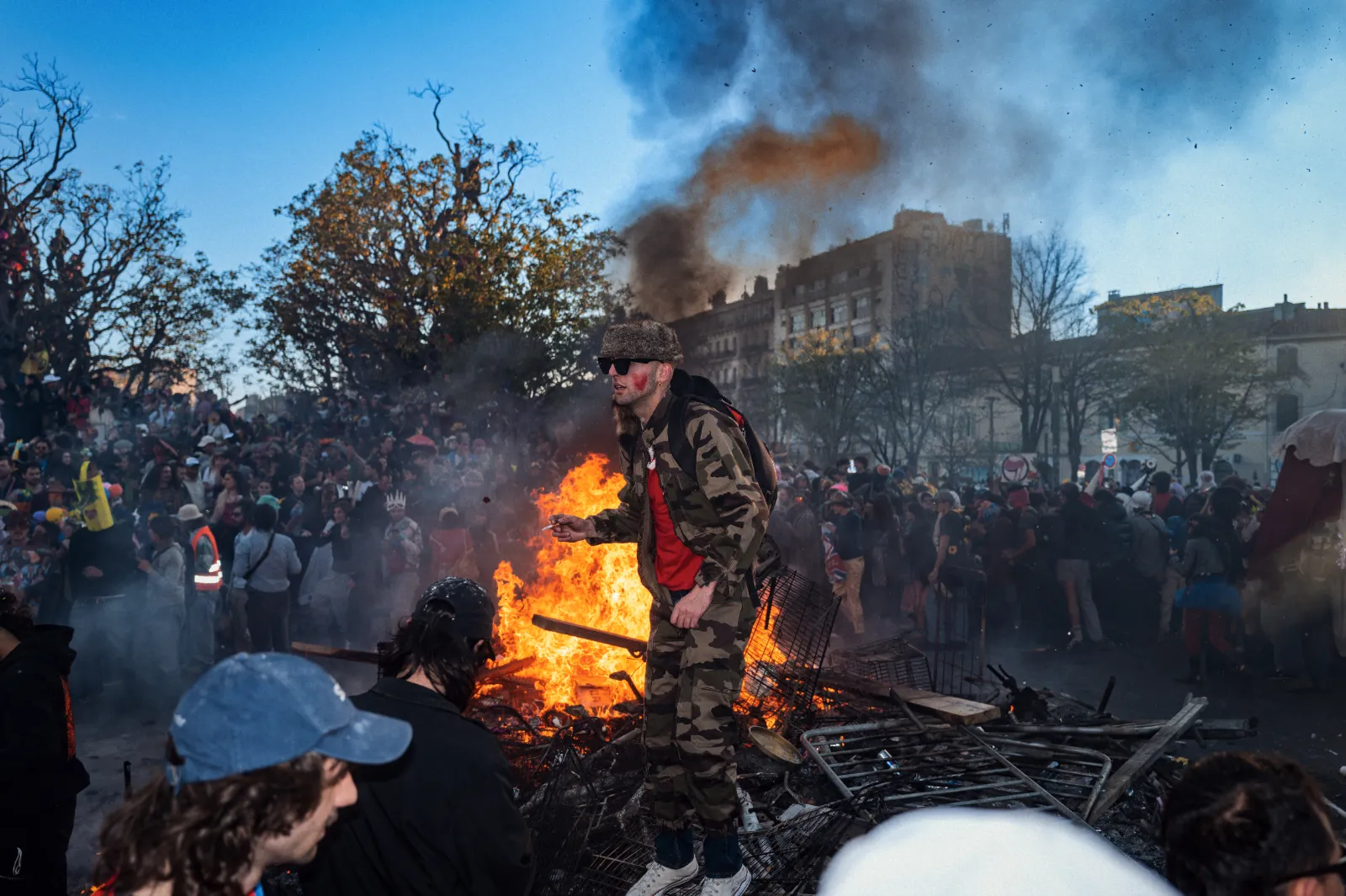Une foule festive autour d'un grand feu de joie, avec des personnes déguisées et un homme en tenue camouflage sur les débris.