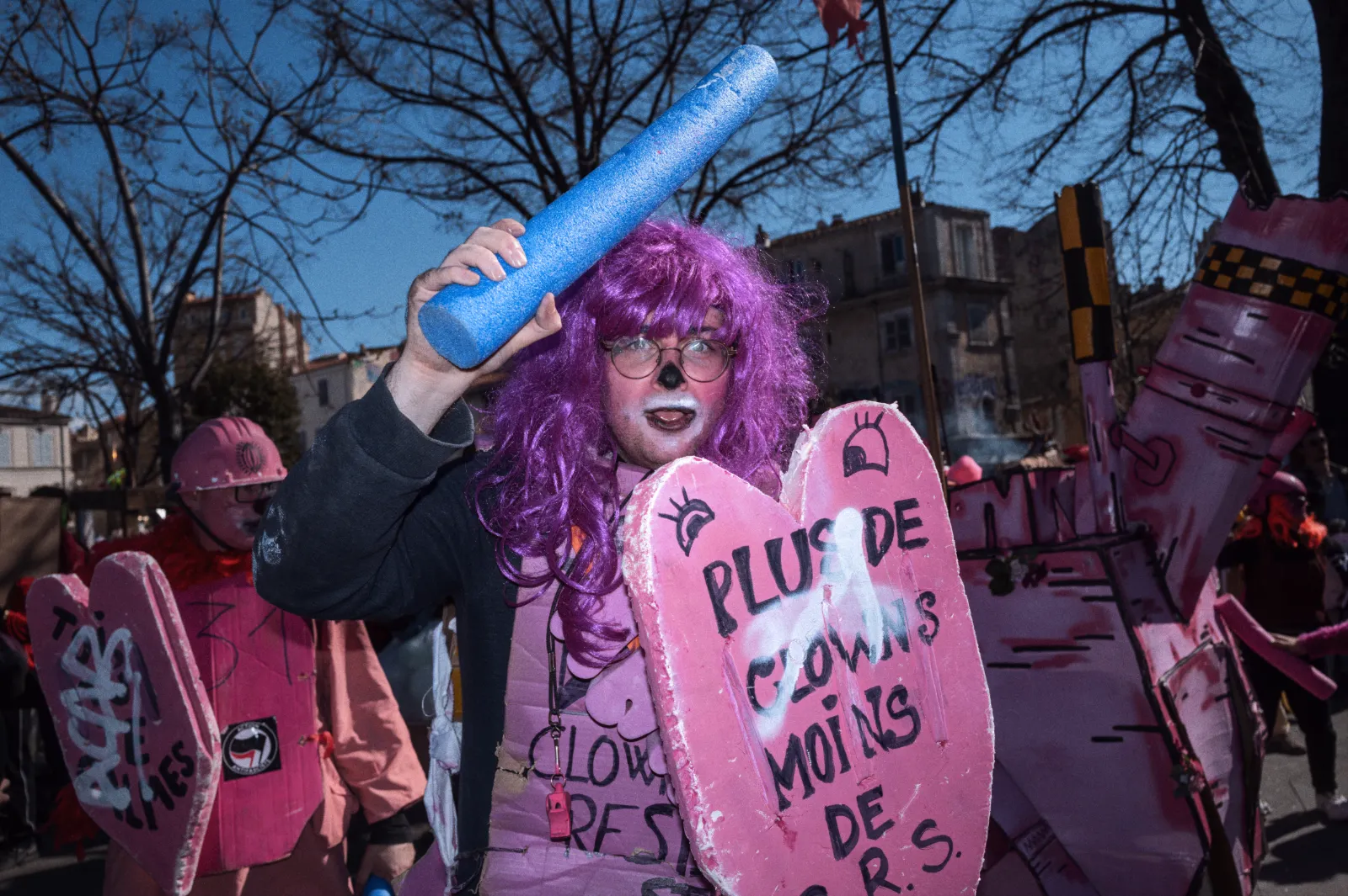 Un participant au carnaval, vêtu d'une perruque rose et portant un bouclier décoré, s'amuse avec un accessoire bleu.