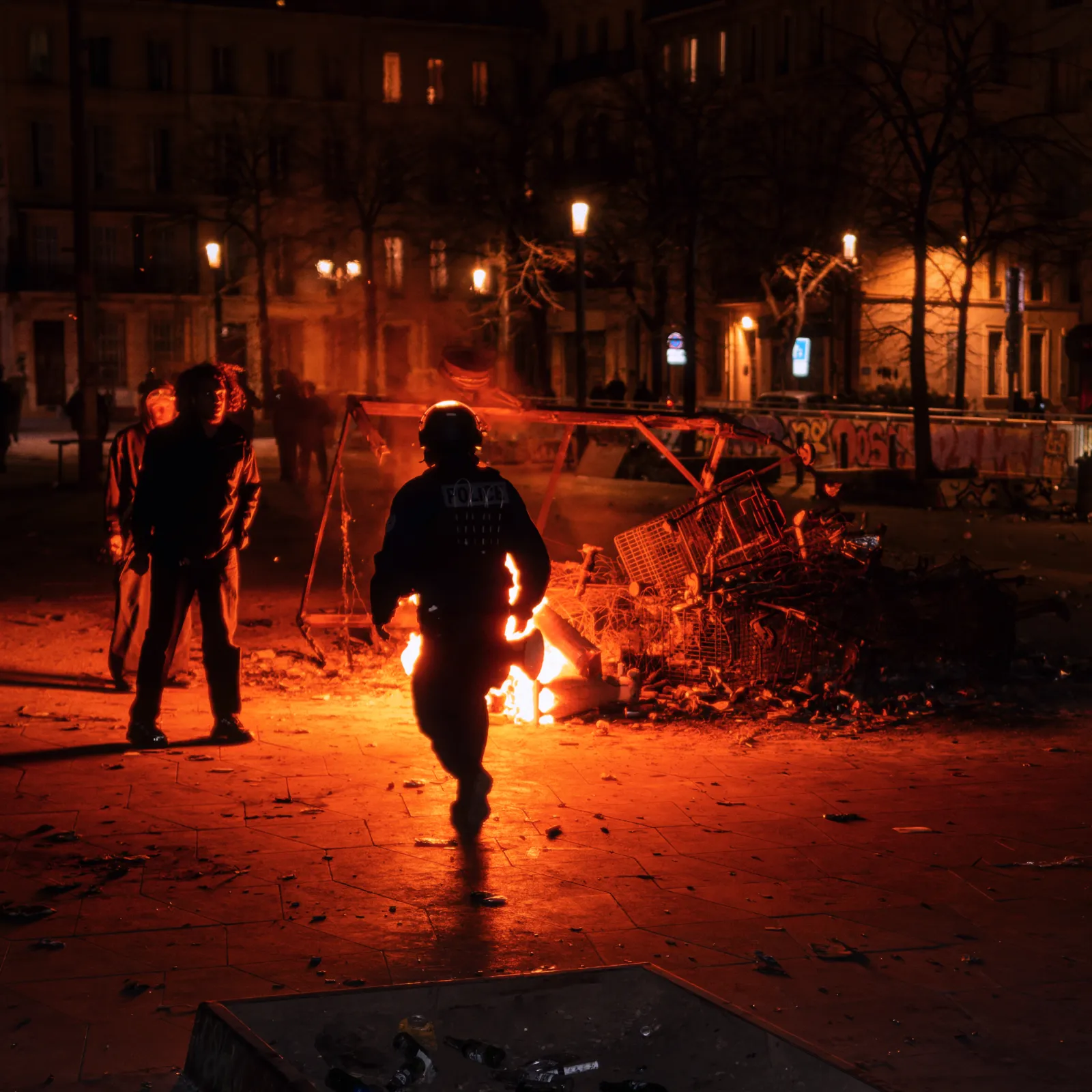 Scène nocturne au Carnaval de la Plaine, Marseille, avec des personnes près d'un feu et des débris en arrière-plan.