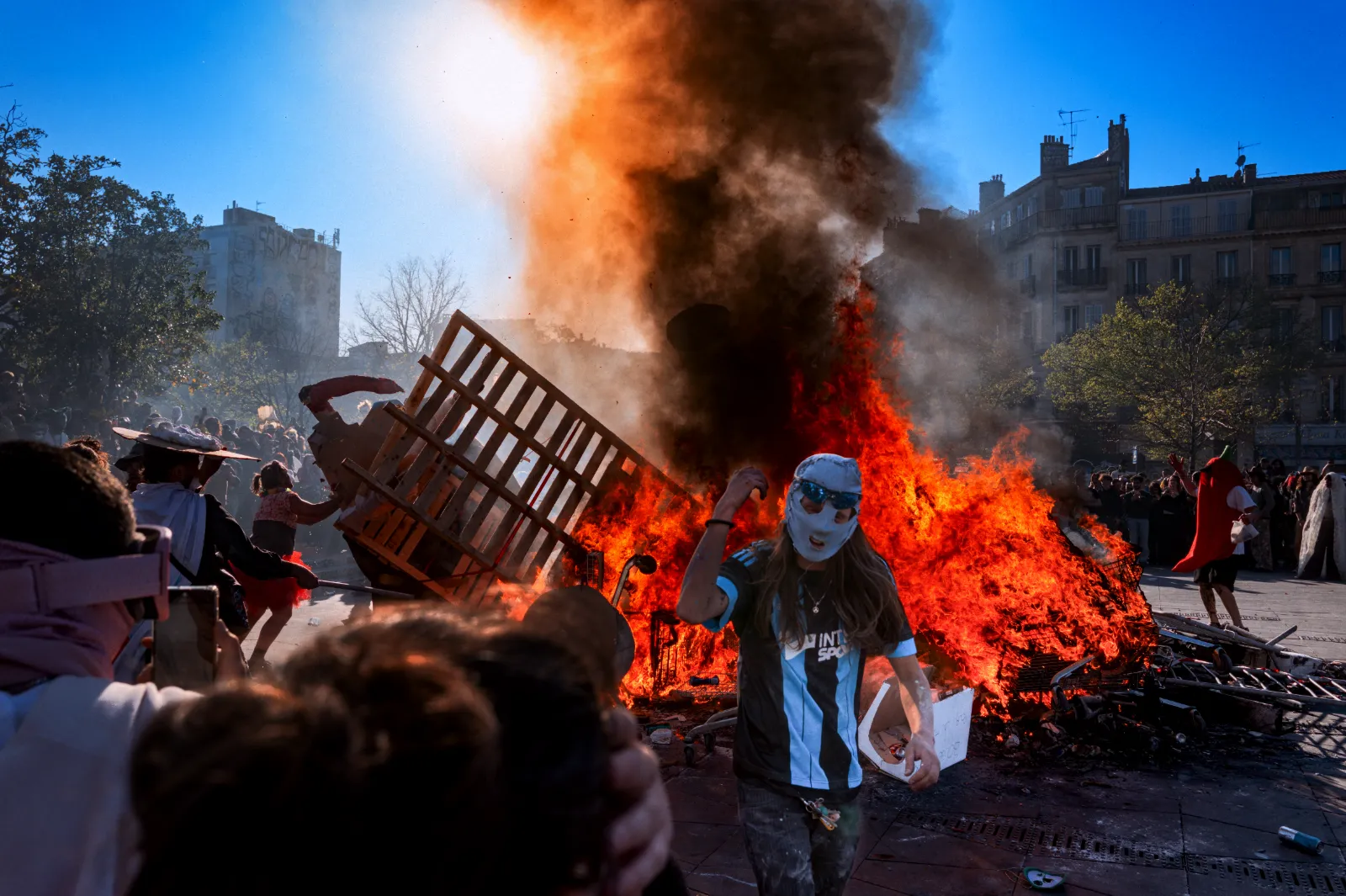 Un groupe de personnes en fête autour d'un grand feu de joie, avec des costumes colorés et des masques, sous un ciel ensoleillé.