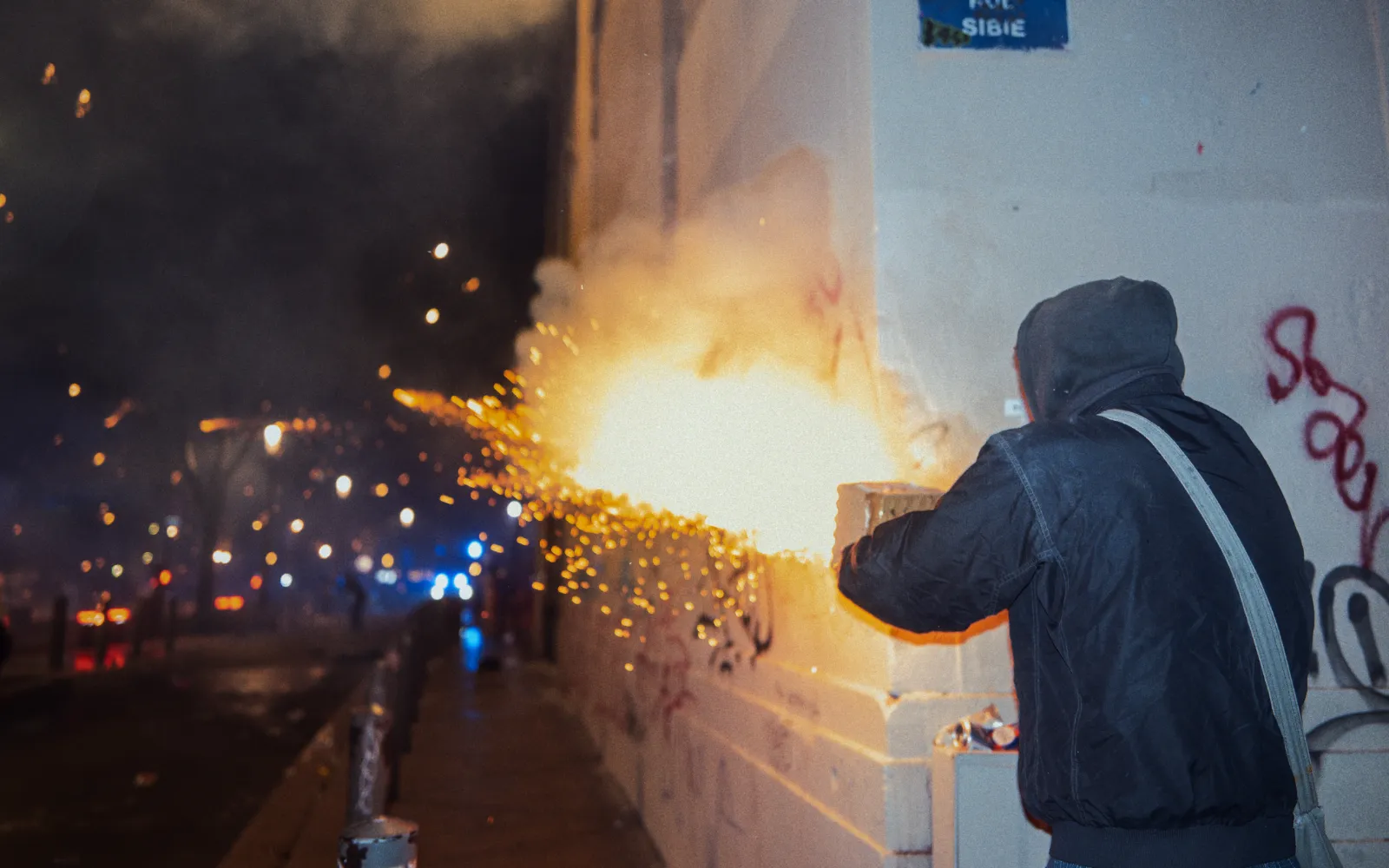 Un participant au carnaval lance des feux d'artifice, créant des étincelles lumineuses dans la nuit. Mur graffiti en arrière-plan.