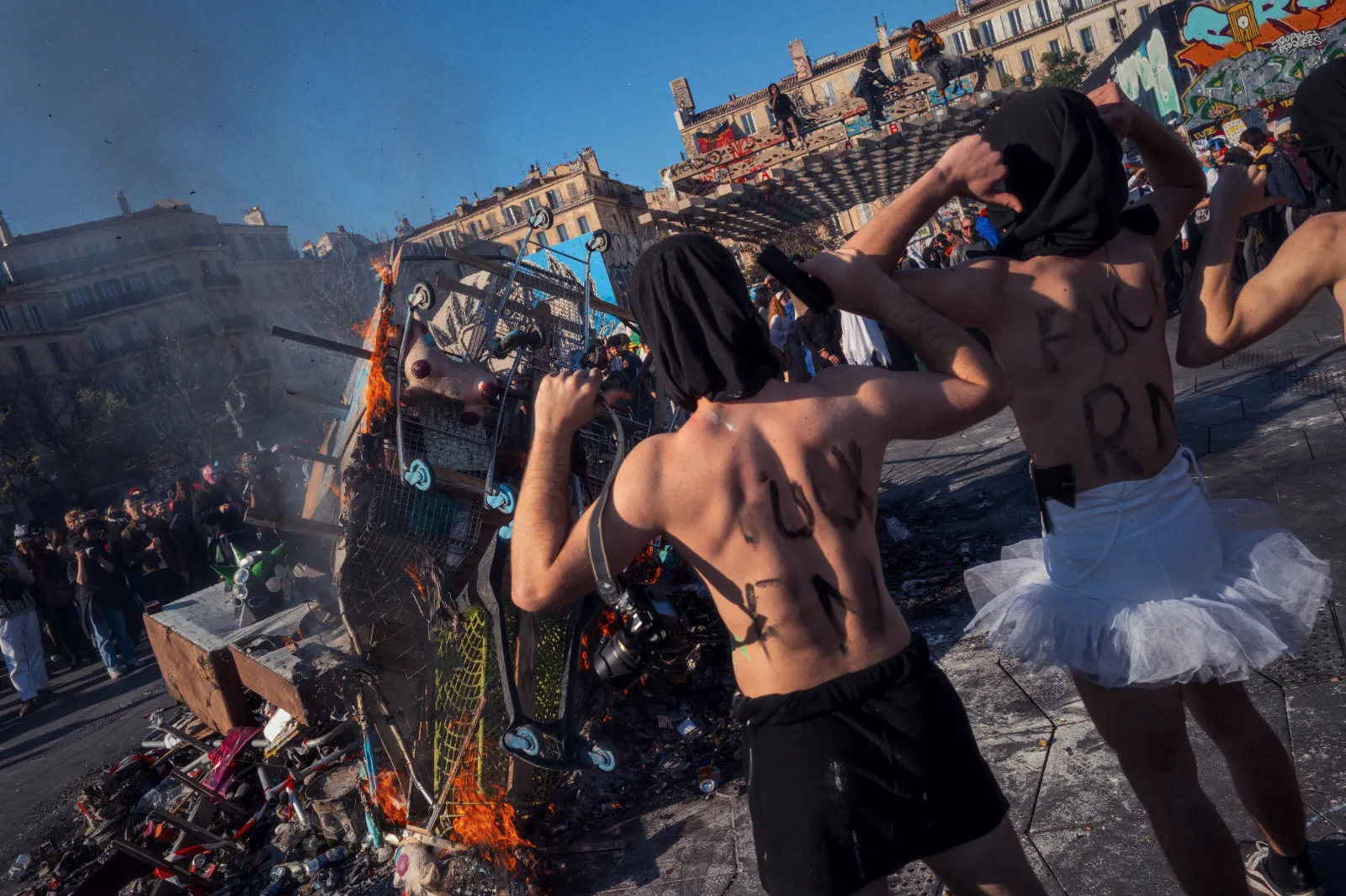 Des participants au carnaval portent des capuches et affichent des messages sur le dos, devant un feu de déchets et une foule festive.