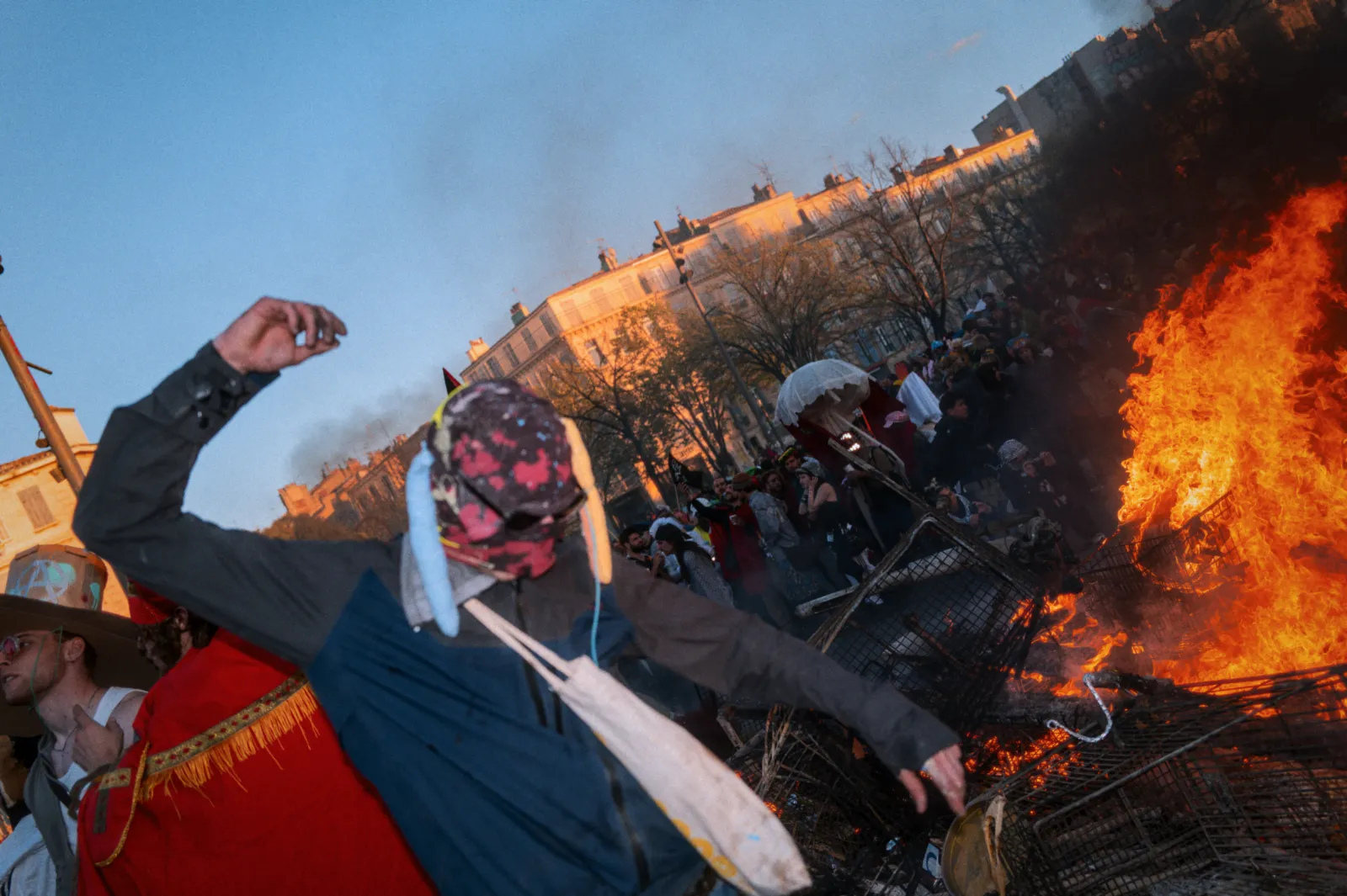Un participant au carnaval danse près d'un grand feu, entouré de spectateurs et de fumée, dans une ambiance festive.