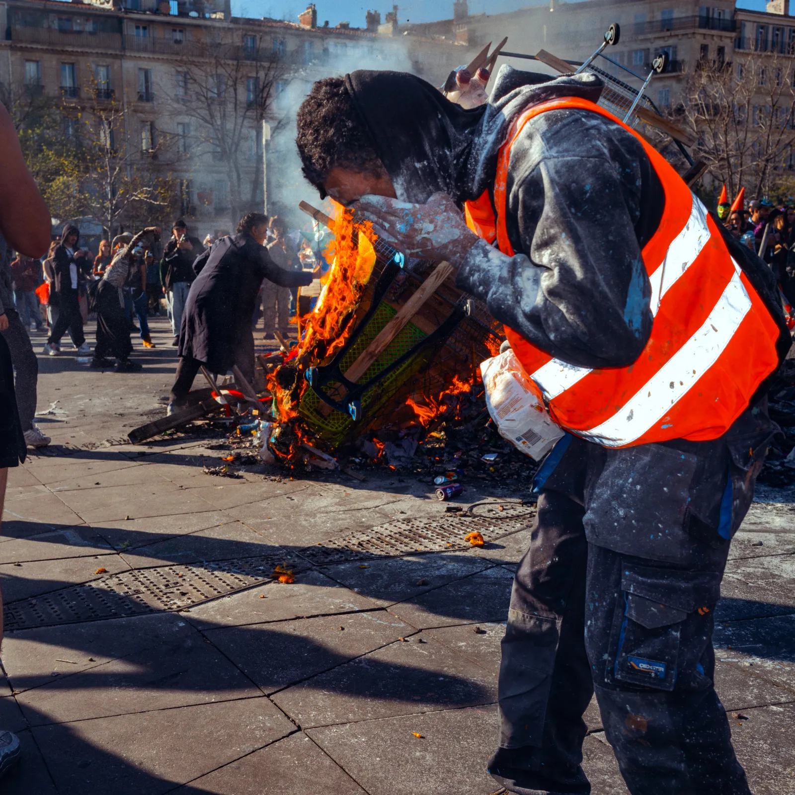 Un homme en gilet orange se penche près d'un feu sur la place Jean-Jaurès, entouré de participants au carnaval.