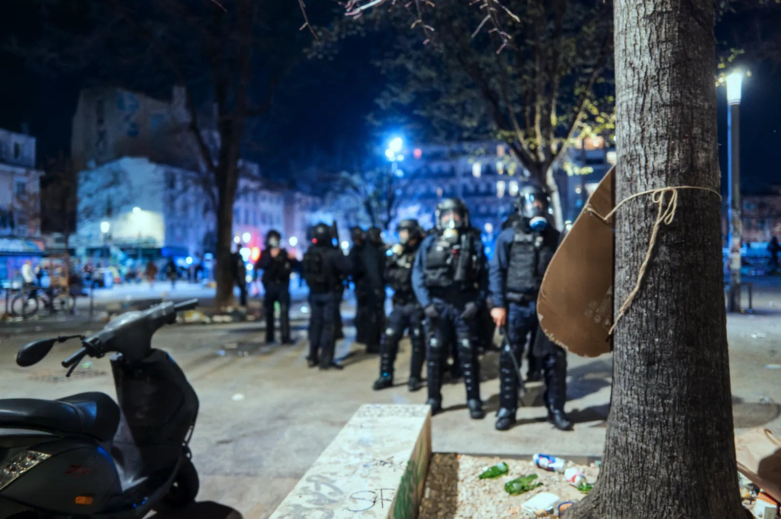 Scène nocturne au Carnaval de la Plaine, avec des policiers en tenue anti-émeute et des déchets au sol près d'un arbre.