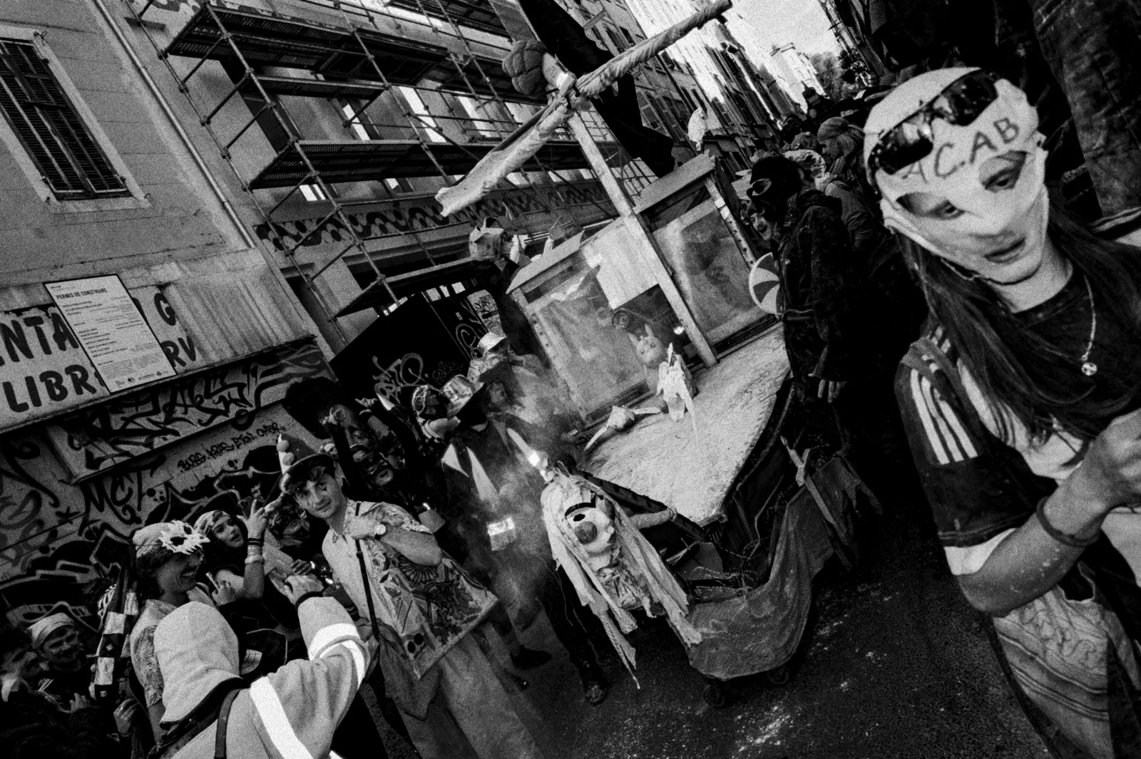Un char décoré défile dans une rue animée, entouré de participants masqués et vêtus de costumes colorés, avec des graffitis en arrière-plan.