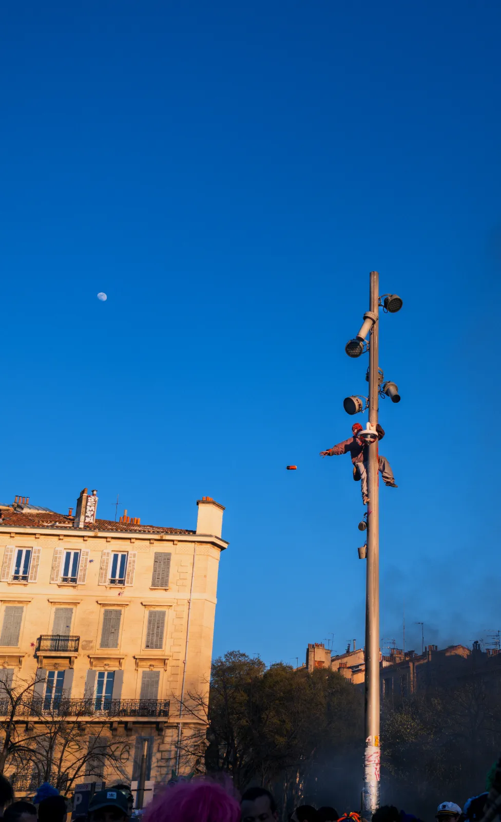 Un lampadaire avec des haut-parleurs, un personnage suspendu, et un ciel bleu clair avec la lune visible. Ambiance festive de carnaval.