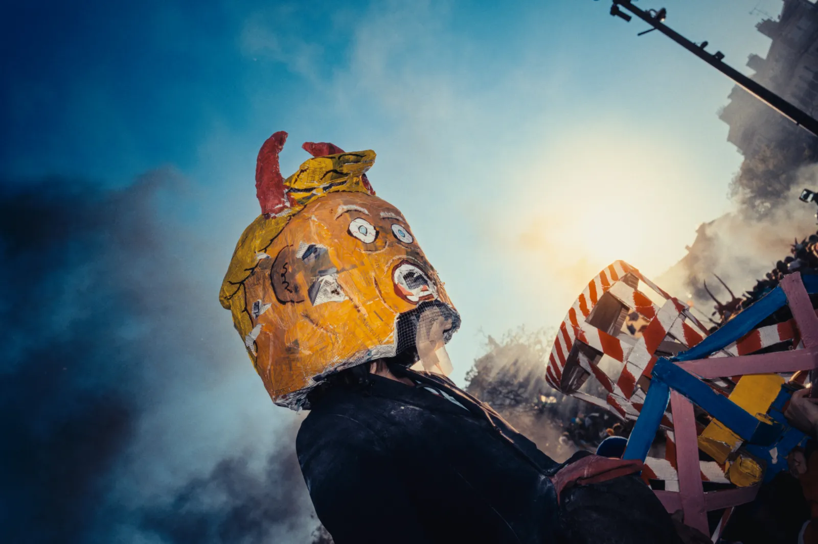 Un personnage masqué avec un grand chapeau coloré, entouré de décorations festives, sous un ciel bleu et ensoleillé.