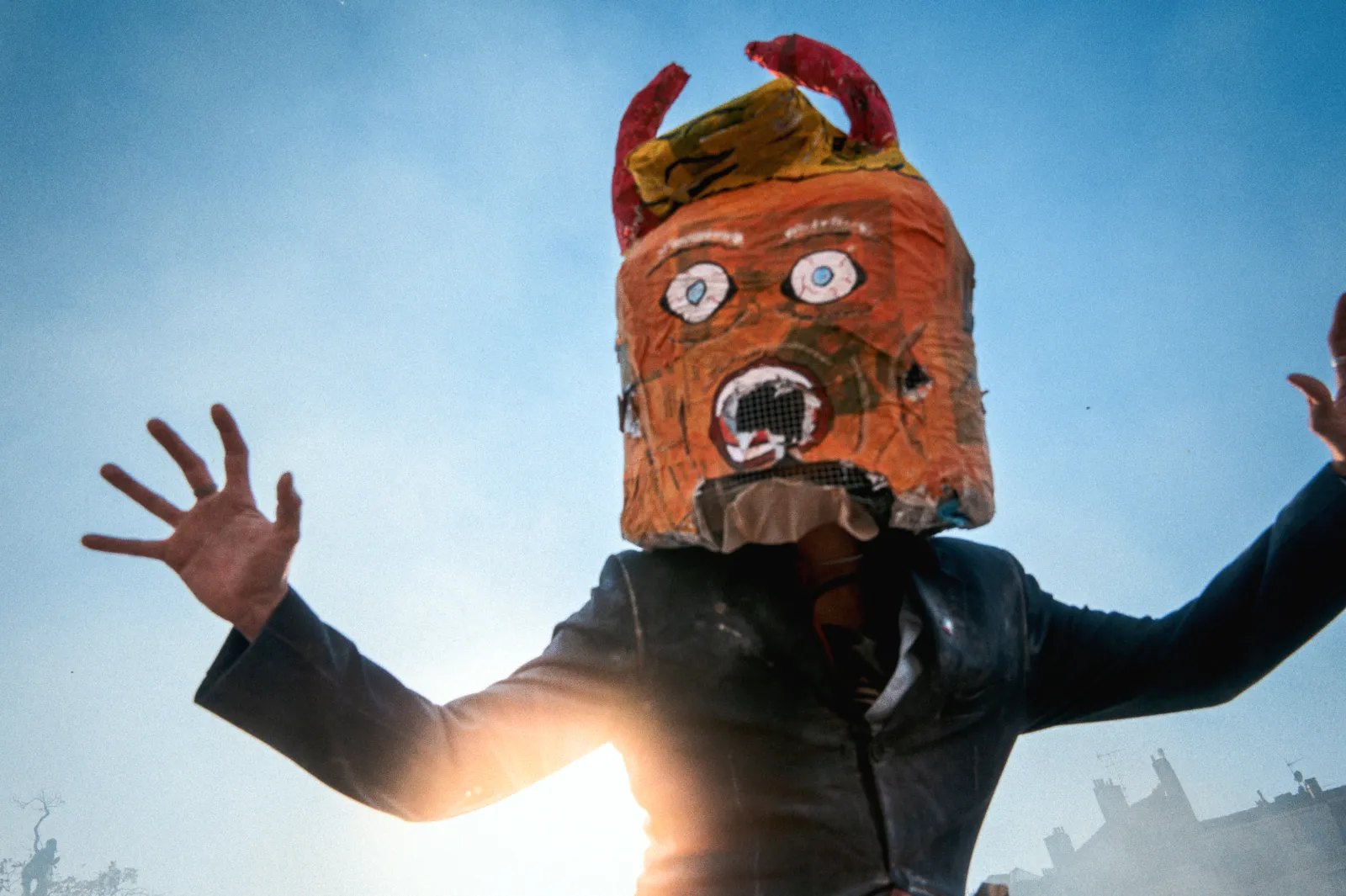 Un participant au carnaval porte un masque en papier mâché avec des cornes, dans une ambiance festive sous un ciel bleu.