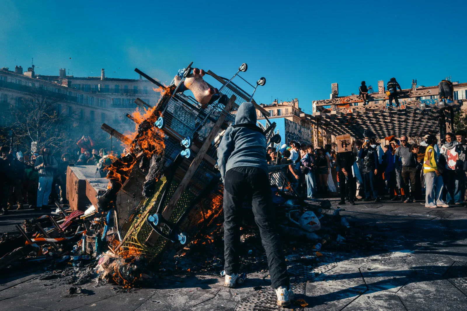 Un homme pousse un grand tas de débris en feu lors du Carnaval de la Plaine, entouré d'une foule sur la place Jean-Jaurès.